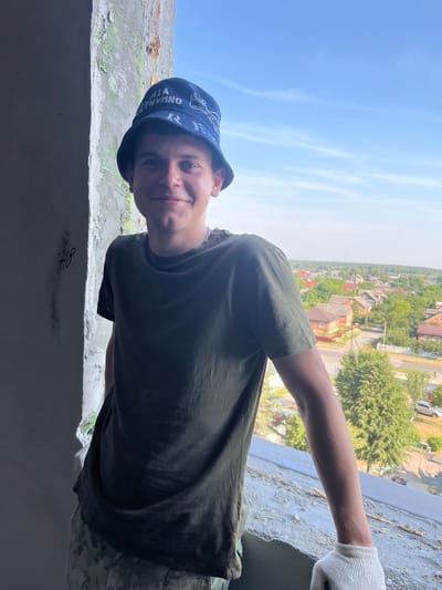 A young man wearing a dark green t-shirt, camouflage pants, and a blue bucket hat stands by an open window, leaning on the windowsill. His left hand is visible, wearing a white work glove. The background shows a clear blue sky and a residential area with houses and trees, indicative of a rural or suburban setting within Аснежыцкі сельскі Савет, Belarus. The window frame appears to be under construction or renovation, with exposed concrete and plaster. The man is looking towards the camera with a slight smile.