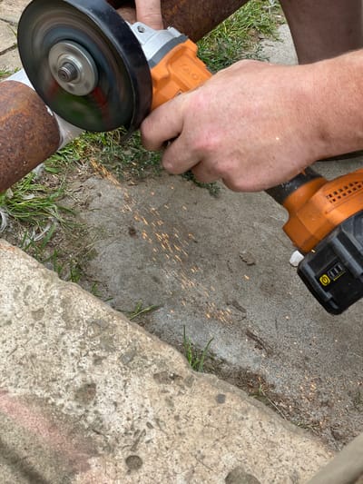 In an outdoor setting, likely on a concrete or dirt surface with patches of grass, a person is using an orange and black angle grinder to cut into a rusty metal pipe. Sparks are flying from the point of contact between the grinding disc and the metal. The person's hand, with visible skin texture and some freckles, is holding the grinder. Part of the person's leg and foot are visible in the background, suggesting they are wearing shorts and possibly work boots or closed-toe shoes. The environment appears unkempt, with some loose debris and vegetation. No specific location in the city of Baldeyka, Russia can be determined from the image.