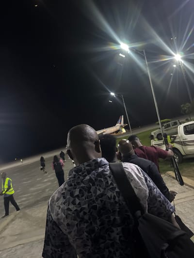 The image depicts a nighttime scene in Omoecheigbo, Nigeria, showing a group of people near an airplane. An individual wearing a patterned shirt and a seatbelt is in the foreground, facing right. Behind them are several other people, including one in a red shirt. Further back, an airplane is visible, with its tail section bearing red, white, and blue colors. Ground vehicles are also visible, including a white van. Streetlights illuminate the scene, casting strong light and glare. A person wearing a yellow safety vest is seen walking on the ground. The sky is dark, and a crescent moon is visible.