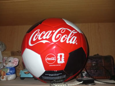 A Coca-Cola branded soccer ball is prominently displayed on a wooden shelf, positioned slightly off-center to the right. The ball is predominantly red with white and black pentagonal panels. The Coca-Cola logo, in white script, is clearly visible across the top of the ball. Below the logo, there are smaller icons and text, including "Coca-Cola", a FIFA logo, and the words "WORLDWIDE PARTNER".
To the left of the soccer ball, a small, ceramic elephant figurine dressed in a blue jacket and holding a green object is visible. Behind the elephant, a portion of a light gray statue or figurine can be seen. Beneath the soccer ball, a small blue toy car with black wheels is partially obscured.
Further to the right of the soccer ball, a dark, intricately carved wooden box with metal accents sits on the shelf. A black cable or wire extends from this box. A portion of what appears to be a white woven mat or placemat is visible in the foreground, partially covered by a black object, possibly a remote control.
The background consists of a wooden shelf, with the wood grain visible. The lighting appears to be ambient, casting subtle shadows. The scene is static and appears to be an indoor display. The image does not contain any elements that would indicate a specific location in the city of Termez, Uzbekistan.