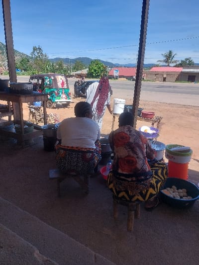 The image depicts an outdoor food stall scene in Kimamba, Tanzania, under a clear blue sky. In the immediate foreground, two women are seated on low wooden stools, their backs to the viewer. The woman on the left wears a white top and a dark patterned skirt with circular motifs. The woman on the right wears a floral top with orange and red patterns and a skirt with a yellow and black chevron-like design. Between and around them are various cooking utensils, including pots, basins, and a metal strainer. A dark green basin to the right of the right woman contains numerous light-colored, spherical food items. A red plastic basin holds a clear bottle of water.
In the mid-ground, a third woman stands facing away, wearing a striped garment and a dark head covering, positioned near a white plastic bucket on a low stand. To her left, a wooden table holds additional cooking vessels. Beyond the stall area, an unpaved shoulder transitions to a paved road. A colorful green three-wheeled auto-rickshaw (tuk-tuk) with decorative images is parked on the roadside, followed by a dark sedan. In the background, sparse trees, single-story buildings with red roofs, and distant mountains are visible under the bright sunlight. A dark wooden support post for an overhang frames the left side of the foreground.