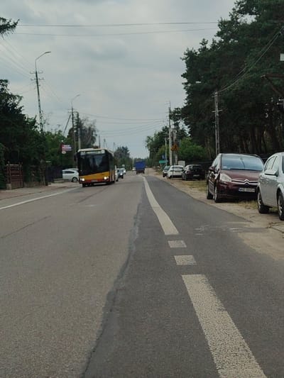 The image depicts a street scene in Łomna, Poland, on an overcast day. A yellow and black public bus, identified by the license plate "WB 8770L", is the primary subject in the foreground, driving on the right side of the asphalt road. The bus has its headlights on. Several cars are parked on the right side of the road, including a maroon Citroën with the license plate "WND 88904". A silver sedan is parked next to it. A dark blue truck is visible further down the road. The street is lined with trees on both sides, with power lines and poles running overhead. A speed limit sign is visible on the left side of the road, near a white building. The road has clear white lane markings. The general environment suggests a suburban or rural area.