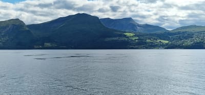 A wide-angle, landscape shot captures a serene body of water in the foreground, reflecting the overcast sky. In the midground, lush green mountains rise dramatically, dotted with scattered buildings and farmland. The distant background is dominated by more mountainous terrain, shrouded in a mix of clouds and sunlight. The scene is likely Volda, Norway, given the distinctive fjords and mountainous landscape characteristic of the region. No people or animals are clearly visible.