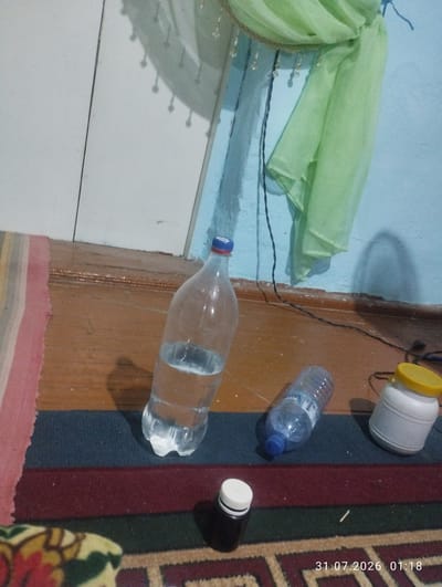 The image shows a scene indoors, likely a room. On a wooden floor, a large clear plastic bottle filled with water is placed. To its right, a smaller clear plastic bottle with a blue cap is visible, and further to the right, a white plastic container with a yellow lid is present. In the foreground, a small dark bottle with a white cap sits on a carpet. In the background, a light blue wall meets a wooden wall panel. A piece of light green fabric is draped on the wall. A date and time stamp, "31.07.2026 01:18", is visible in the bottom right corner. The location is identified as Quyosh, Uzbekistan.