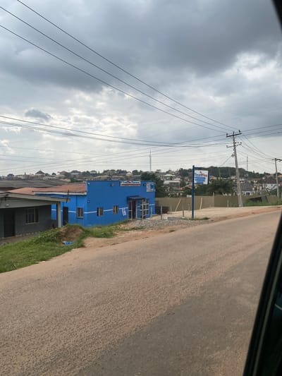 The image shows a street scene in Ojere, Nigeria, under a cloudy sky. In the foreground, a paved road curves to the right. To the left of the road, a bright blue building with a red roof stands out. A sign above the building's entrance reads "TrustPlus Pharmacy". Next to the blue building, a smaller, grey building with a similar red roof is partially visible. Beyond these buildings, a residential area with houses and trees covers a hillside, with power lines stretching across the sky. Several utility poles are also present, supporting the overhead wires. The overall impression is of a developing urban area with commercial and residential structures.