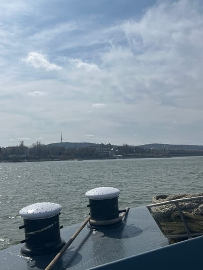 The image captures a view from a vessel's deck across a body of water towards the city of Cuza Vodă, Romania. In the foreground, two dark-colored bollards with white, studded, domed tops are affixed to a grey, textured deck. A light-colored rope is positioned near the base of the right bollard. To the far right, coiled ropes are contained within a metal railing structure. The midground shows a wide river with rippled water reflecting sunlight. The distant background features the urban profile of Cuza Vodă, characterized by a prominent, slender telecommunication tower situated on a hill. Bare trees line the far riverbank, with multiple buildings visible, including a multi-story structure with a distinctive roof on the left. The sky is partially cloudy, displaying white and grey cloud formations against patches of lighter blue. No individuals are visible.