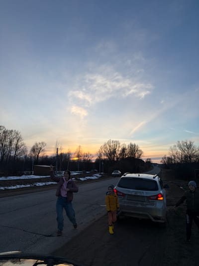 The image shows a road in Дулесово, Russia, at sunset. A silver car is parked on the side of the road. Three people are present: a person in a pink jacket and jeans stands on the road, a child in a yellow jacket and orange pants stands near the rear of the car, and another person in a dark jacket stands further away from the car. Trees are visible along the roadside, silhouetted against the colorful sky. Patches of snow are on the side of the road.