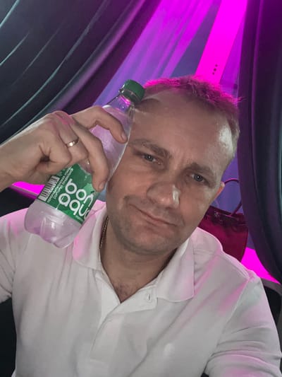 This is a selfie of a Caucasian man with short, light brown hair and blue eyes, looking directly at the camera with a slight smile. He is wearing a white collared shirt. In his right hand, he holds a clear plastic bottle of water with a green cap. The label on the bottle is primarily green and white with text that appears to be in Cyrillic script. A ring is visible on his left ring finger.
The background is dark and indistinct, with vibrant pink and purple lighting. There appear to be draped fabrics or curtains in shades of dark purple and black, with streaks of bright pink light shining through. The lighting suggests an indoor setting, possibly a venue with stage lighting or an event space. The overall mood is casual and relaxed. The visible text on the bottle reads "Bon Aqua" in large, stylized letters, and below that, "ПЕРЕДАТИВНАЯ ВЛАГА УПАКОВОЧНОЙ" which translates to "TRANSFER MOISTURE PACKAGING." The barcode is also visible. Given the location context of Orsha, Belarus, it is likely the man is in this region.