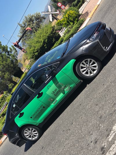 A black Toyota sedan with a distinctive green Uber Eats livery is parked on the side of a street in Collado Villalba, Spain. The car's side is predominantly covered in a bright green vinyl wrap featuring the Uber Eats logo, a stylized coffee cup, and Spanish text that reads "Descarga la app y haz tu pedido" (Download the app and place your order). The vehicle is positioned at an angle, with its front facing towards the right side of the frame. The street is paved with asphalt and shows some white line markings.
In the background, lush green trees and bushes are visible, indicating a somewhat natural or park-like setting. Beyond the vegetation, a hilly landscape with some scattered buildings and structures can be seen. A prominent feature in the upper left of the image is a large, red and black industrial-looking pipe structure mounted on a pole, possibly part of a water or irrigation system. Overhead, power lines or cables crisscross the sky, and a clear blue sky with bright sunlight suggests a warm day. A few road signs are also present in the upper right portion of the image, including a directional sign that appears to read "EL BERROCAL". The overall scene suggests an outdoor urban or suburban environment with a mix of man-made structures and natural elements.