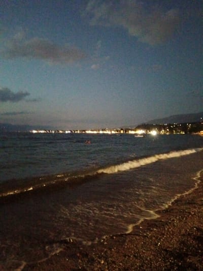 The image depicts a nighttime view of a beach along the Municipal Unit of Vrachneika, Greece. The sky is dark blue with scattered, soft clouds. The sea is calm with small waves gently lapping onto a pebble beach. In the distance, a coastal town is illuminated by numerous lights, creating a blurred line of brightness against the darker silhouette of hills or mountains. A few faint lights are visible on the water, possibly from boats. The overall scene is tranquil and atmospheric, characterized by the soft glow of the distant town and the subtle interplay of light on the water and shore.