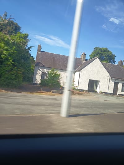 The image shows a view of a white-painted house with a dark-tiled roof, set against a blue sky with some wispy clouds. The house appears to be a single-story or bungalow-style dwelling with multiple windows and doorways. A tall, white vertical pole runs through the center of the frame, obscuring a portion of the house and creating a blurred effect. Green trees and bushes are visible to the left and behind the house, and a paved driveway or area is in front. The foreground shows a blurred view of a road. This appears to be a residential property, possibly located in a rural or suburban area of Killarney, Ireland, given the context of the request. There are no people visible in the image.