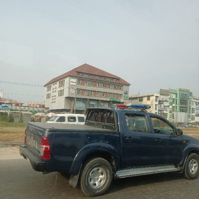 The image, rotated 90 degrees clockwise from its natural orientation, captures an outdoor urban street scene in Nigeria during daylight hours. The foreground is dominated by a dark blue Toyota Hilux pickup truck, viewed from its side and rear, positioned on the right side of a paved road. The truck is equipped with a metallic side step bar, a covered bed, and a distinctive red and blue emergency light bar mounted on its roof, suggesting it is an official or security vehicle. The rear tailgate clearly displays the "TOYOTA" logo, with "Hilux" partially visible underneath.
In the mid-ground and background, a series of multi-story buildings line the road. The most prominent structure on the left features a reddish-brown sloped roof and light gray walls with multiple rectangular windows. Several commercial signs are visible on its lower floors, one of which appears to read "Maison." Behind this building and further along the road, other buildings of varying designs, including one with a green facade, contribute to the city landscape. A white vehicle is partially visible behind the blue pickup truck's bed, suggesting other traffic or parked cars.
The sky is bright but appears somewhat hazy, indicating a clear day. No individuals are distinctly visible within the frame. The scene conveys a typical road environment within an urban or suburban area.