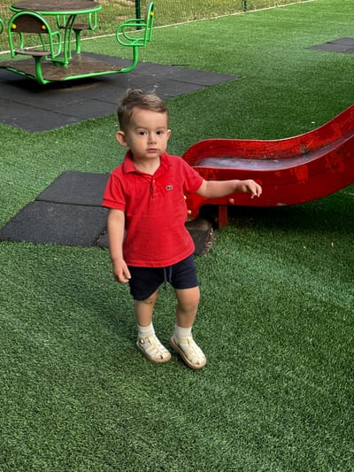 The image shows a young child standing on a bright green artificial turf surface in a playground. The child is wearing a red polo shirt with a small white logo on the left chest, and navy blue shorts. They are also wearing white socks and light-colored sandals. To the left of the child, there is a portion of a playground structure with a circular platform and green frames, possibly a merry-go-round or a similar ride. To the right and behind the child, a portion of a red slide is visible. The background consists of more artificial turf and some fencing or other playground equipment. The overall impression is of a sunny day at a children's play area.