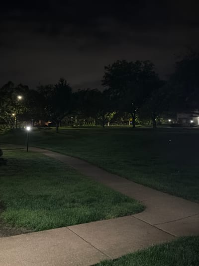 The image depicts a dimly lit outdoor scene at night, likely in a suburban park or residential area in Vernon Hills, United States of America. A paved walkway curves through a grassy lawn, illuminated by a solitary lamp post to its left. Silhouetted trees are scattered across the background, with a building faintly visible on the right side of the frame. The sky is dark and overcast, with no visible stars or moon. The overall atmosphere is quiet and serene.