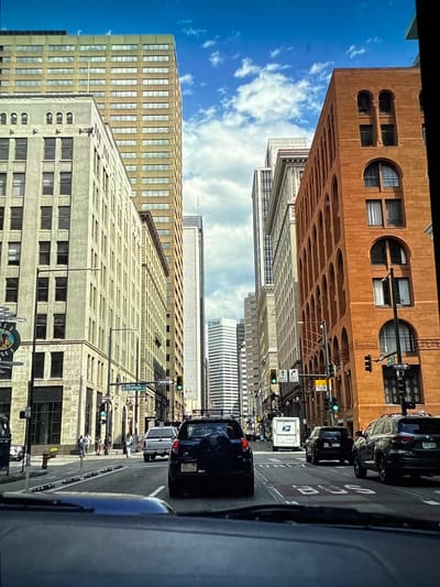 The image shows a street scene in Denver, United States, viewed from a vehicle. The sky is bright blue with scattered white clouds, suggesting it is daytime. The buildings lining the street are tall and of various architectural styles, some with brick facades and intricate window designs, others with more modern, streamlined appearances. Cars are present on the road, indicating traffic. One car has a license plate that reads "GGN 672". There is a street sign that says "Champa". The overall impression is a busy urban environment under clear weather conditions. There are no discernible people engaged in specific activities, nor any clear indications of emotions. The focus is on the architecture and the urban infrastructure.
