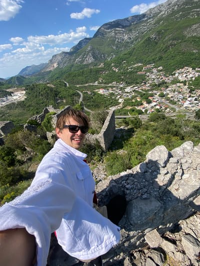 The image depicts a selfie taken from a high vantage point, likely on a rocky mountain path or overlook in Zgrade, Montenegro. In the foreground, positioned to the right, a young man with light brown hair and dark sunglasses is smiling directly at the camera. He is wearing a white collared jacket, and his right foot, clad in a dark shoe, is visible on the rocky ground in the lower right corner.

Behind the individual, a breathtaking panoramic view unfolds under a bright, clear blue sky with scattered white clouds. A steep, verdant mountainside, densely covered with trees, dominates the midground. Nestled within this green landscape is a town or village characterized by numerous white and light-colored buildings. Winding roads are visible, snaking through the terrain and connecting various parts of the settlement. In the far distance, additional layers of rugged grey mountains rise. The strong sunlight casts distinct shadows on the rocks, suggesting a clear day, likely late morning or early afternoon. The scene conveys a sense of adventure and enjoyment of the majestic natural and human-built environment. No visible text is present.