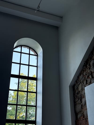 This image depicts the interior of a building with a large, arched window featuring multiple panes and a dark frame. Through the window, lush green trees and a bright sky are visible. The interior walls are painted white, with a section of rough, stone and brick wall on the right side of the frame. A modern track lighting fixture is mounted on the dark ceiling. A small circular security camera is visible on the angled section of the wall on the right. The overall impression is one of a somewhat historic or rustic interior space with a modern aesthetic. The location is identified as Ilyanskiy selskiy Sovet, Belarus.