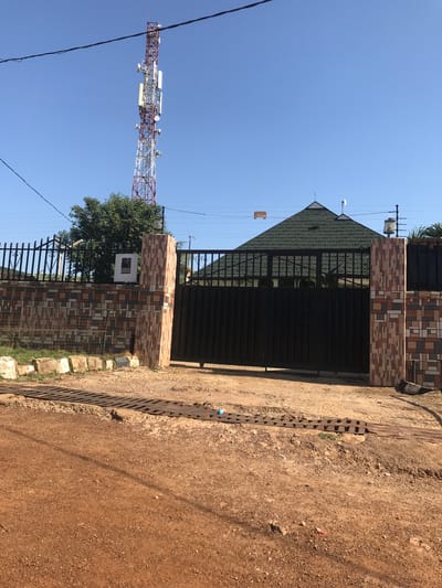 The image displays an outdoor scene featuring the entrance to a property, a dirt road, and a prominent telecommunications tower under a clear blue sky.
In the center, a dark metal gate serves as the entrance, flanked by sturdy gate posts decorated with a mosaic of multi-toned tiles in shades of brown, beige, and white. Behind the gate, a building with a dark green, pitched roof covered in tiles is partially visible. A black metal picket fence extends from the gate, with a verdant tree growing behind it to the left. An outdoor light fixture is mounted above the gate, and another is on the left gate post.
To the far left, a tall telecommunications tower dominates the skyline, characterized by its red and white lattice structure and numerous antennas and dishes. Utility lines crisscross the upper part of the frame, with a small, light-colored rectangular object (possibly cardboard) hanging from one.
The foreground consists of an unpaved, reddish-brown dirt or gravel road. A metal drainage channel or grate runs along the road's edge, adjacent to the property. On the right gate post, a white box bears a black and white sign that clearly reads "NOTICE" at the top, with additional text below that appears to be "PROPERTY" or "PREMISES."
The scene is well-lit by bright sunlight, indicating daytime with clear weather. No people or active events are present, conveying a static view of a residential or commercial area in what appears to be a developing region. The location context is Mandate Estate, Ilorin, Nigeria.