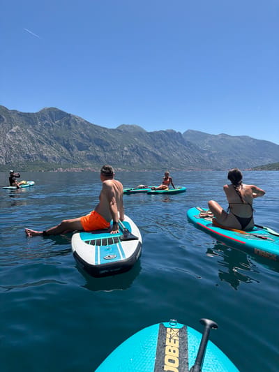The image shows a group of people on paddleboards on the water near Dražin Vrt, Montenegro. The sky is blue, and large mountains are visible in the background. One man is standing on a paddleboard wearing orange shorts. A woman in a black bikini is kneeling on a paddleboard. Several other paddleboarders are in the distance. A paddleboard with "JOBE" written on it is visible in the foreground.