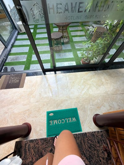 A first-person perspective shot captures a view from inside a building, looking out through a glass door and window. The scene shows an outdoor patio area with artificial turf interspersed with large, light-colored stone tiles. There are several woven furniture pieces, including a table and two chairs, as well as potted plants. A name or logo, "HEAVEN HOME", is visible etched onto the glass, along with a phone number. Inside the building, near the entrance, a brown doormat with the word "HELLO" is placed on the floor. Further into the interior, a green doormat with the word "WELCOME" is positioned. The floor inside appears to be tiled with large, beige, marble-patterned tiles. A person's legs and feet are visible in the foreground, with one foot bare and wearing a black toenail polish, the other foot partially visible. The person is wearing a light pink garment. Dark brown, cylindrical railings are also present. The location is identified as Cam Lâm, Vietnam.