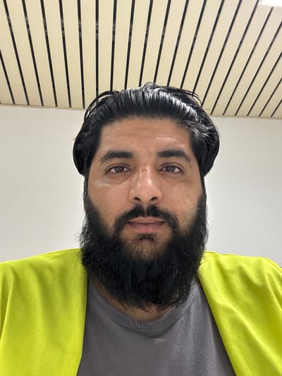 A close-up, head-on, eye-level, full-face photograph captures a man with dark, slicked-back hair and a full beard. He is wearing a bright yellow, long-sleeved garment over a gray t-shirt. The background consists of a white wall and a ceiling with parallel, cream-colored slats separated by black lines. The lighting is bright and uniform, casting minimal shadows. The image appears to have been taken indoors. There are no discernable features that would indicate the location within the city of Schweitenkirchen, Germany.