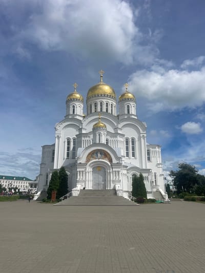 The photograph captures a frontal view of the Holy Trinity Cathedral in Diveyevo, Russia, a prominent religious structure with white walls and golden domes. The cathedral is situated in a vast, paved plaza, and a wide set of stairs leads up to its main entrance. To the left of the cathedral, a building with a green roof and multiple windows is visible, along with some trees and lampposts. Several people are scattered throughout the plaza and near the green-roofed building, some walking and others standing. The sky above is a mix of blue and white clouds, indicating a daytime scene. The overall environment is well-maintained and appears to be a public space.