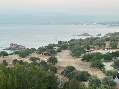 This is an elevated, wide-angle, medium-shot, outdoor, daytime photograph of a coastal landscape with a view of the sea and a settlement. The sky is overcast and a light haze obscures the distant coastline. In the foreground, a dense cluster of green trees occupies the bottom half of the image.
Moving into the mid-ground, a clearing is visible, sparsely populated with trees and dotted with several small, triangular-roofed wooden structures, reminiscent of cabins or bungalows. Some are positioned in a cluster, while others are more spread out. A dirt road winds through this area, and several cars are parked near the cabins.
Further back, a larger open-air structure with a covered roof is set up, appearing to be an event space with tables and string lights. A few people are visible around this structure, and there are other scattered figures and objects in the clearing, including a children's play structure with red and yellow elements.
The background features the calm, greyish-blue sea, stretching to the horizon. A white boat with a dark stripe is visible on the water, and a few distant, low-lying landmasses and the faint outlines of buildings suggest a coastline in the far distance. Rocky outcrops are present in the sea closer to the shore. The overall scene depicts a tranquil, somewhat remote, coastal resort area, possibly within or near Polis Chrysochous, Cyprus, given the typical landscape of the region.