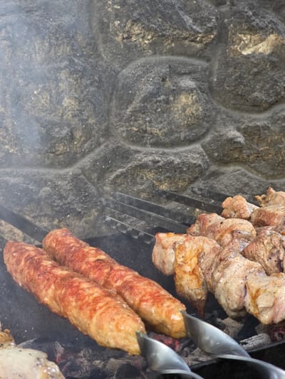 This close-up image shows several skewers of meat cooking over hot coals. The meat on the skewers appears to be a mix of cubed pieces of what looks like pork or lamb, and long, cylindrical sausages. The meat is browned and slightly charred in places, indicating it is in the process of being grilled. Smoke is visible rising from the coals, adding to the atmosphere of an outdoor barbecue.
The background of the image features a rough, textured stone wall, likely part of an outdoor cooking area or fireplace. The stones are varied in color and shape, with visible mortar lines. The overall impression is of a rustic, perhaps traditional, setting for preparing food.
The focus is on the food and the cooking process. There are no people visible in the frame, nor any clear indications of the time of day or specific weather conditions. The lighting suggests it is daytime, and the smoke implies an active cooking session. The location context provided places this scene in Pidhorodne, Ukraine, suggesting a possible cultural or culinary tradition of grilling meat. The image highlights the appetizing appearance of the grilling meat, with the focus on its texture and color. There is no visible text in the image.