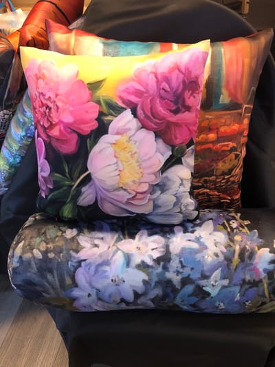 The image shows a close-up of three decorative pillows arranged on a black surface, likely a sofa or chair. The foreground pillow is a long bolster pillow with a dark background covered in a floral print of light purple and blue flowers. Behind it, a square pillow features a vibrant painting of pink and white peonies against a bright yellow background. To the right of that, another square pillow displays a more abstract painting with vertical strokes of orange, red, and teal. The background is dark and out of focus, with some hints of other colorful items. The scene appears to be indoors, possibly in a home or retail setting. There are no people or animals visible. The image does not contain any information to indicate its location in Severn, Canada.