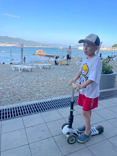 A young boy wearing a white t-shirt with a yellow duck graphic, red shorts, a baseball cap, and grey Crocs rides a three-wheeled scooter on a paved walkway. He is positioned in the foreground, with a beach and the sea in the background. The beach is covered in pebbles, with several white lounge chairs and umbrellas. Several people are visible on the beach and in the water. The sea is calm, with mountains in the distance. The sky is blue with a few scattered clouds. The image appears to have been taken in Ivanovskaya, Russia, based on the provided location context.