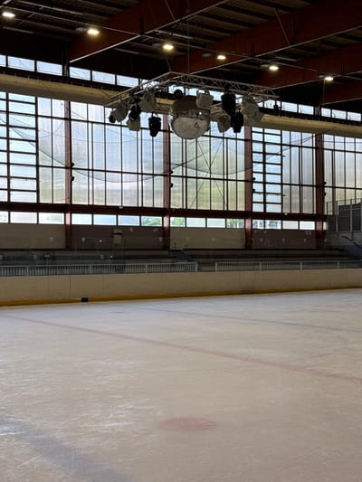 The image shows an ice rink in Garges-lès-Gonesse, France. The ice surface, marked with lines, occupies the right half of the frame. The left side features a wall with large windows and a metal structure supporting lighting equipment, including spotlights and a disco ball. The building's interior has a reddish-brown ceiling. No people are visible.