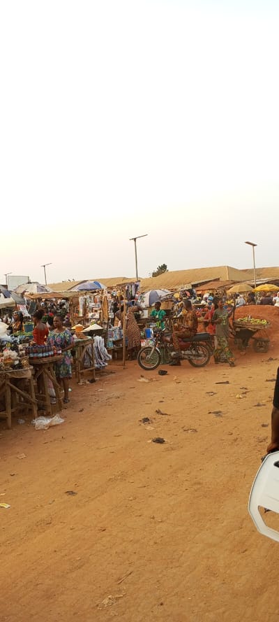A bustling outdoor market scene under a pale sky. Vendors display goods from wooden stalls and carts. People gather, some in colorful patterned dresses. A motorcycle sits amidst the activity. The ground is dusty and worn. Overhead, solar-powered streetlights stand tall. Roofs of nearby buildings peek from behind the crowded stalls.