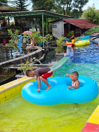 Two young boys are riding a large, blue, double-seated inflatable tube down a water slide in a water park in Kecamatan Tulung, Indonesia. The boys are wearing swimsuits and the younger boy has goggles on. The slide is made of concrete and is lined with yellow and black stripes. The water in the slide is a bright green color and there are some small waves in the water. There is a green roofed structure behind the boys, along with trees and other vegetation. There are also other people in the water park.