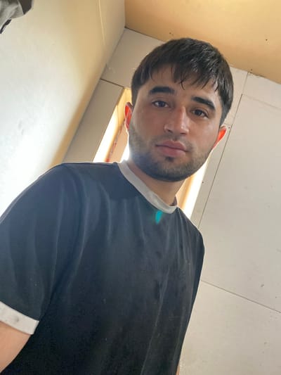 A medium shot is captured from a low angle, looking up at a young man with dark hair, dark eyes, and a short beard. He is wearing a black t-shirt with white trim around the collar and sleeves. The man is positioned in the foreground, with his face slightly tilted and looking towards the camera. The background consists of light-colored walls, a ceiling, and what appears to be a window or opening emitting bright light, creating a high-contrast scene. The environment suggests an indoor setting, possibly unfinished or under construction, with visible seams in the wall panels and some dust particles on the surfaces. The image does not provide any information to determine the location in the city of Vesniny, Russia.