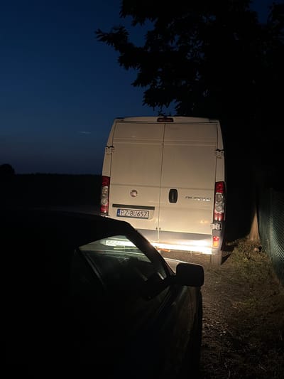 In Rosnówko, Poland, at night, a white Fiat Ducato van is parked on a dirt road next to a dark-colored car. The van's brake lights and reverse lights are illuminated. A license plate on the van reads "PZ 8U657". A chain-link fence is visible to the right of the van, and a tree casts a shadow over the scene from the upper right. The sky is dark blue, indicating twilight or early night. The ground appears to be covered in dirt and dry vegetation.