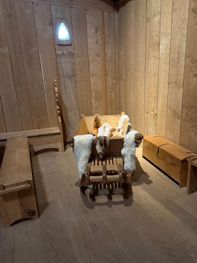 The media file shows an interior shot of a room with wooden walls and floor, characteristic of a rustic or traditional setting. The room is sparsely furnished with wooden objects, including shelves and what appears to be a decorative display. In the center of the image, there is a display featuring animal furs, possibly for decorative or historical purposes. A small, triangular light fixture is embedded in the wooden wall on the left side of the frame, emitting a bright white light. The overall impression is one of a quiet, possibly historical, space. There are no people visible in the media file, and no text is present. The lighting suggests an indoor environment, possibly lit by artificial sources. The wooden interior suggests a location that values natural materials and traditional craftsmanship.