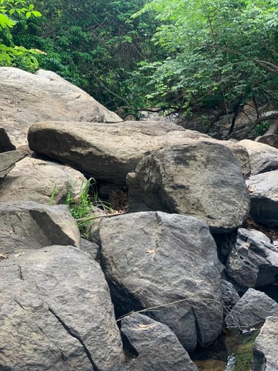 The image shows a close-up view of a rocky area in Egbeda, Nigeria. The location is near a stream or drainage channel. The rocks are large, gray, and weathered. Some of the rocks have moss growing on them. A few small plants grow in the crevices between the rocks. The image was taken from a low angle, looking up at the rocks. A small patch of green foliage can be seen in the top left corner, while a small section of blue sky is visible in the upper right. There are no people in the image. The image was likely taken during the day.