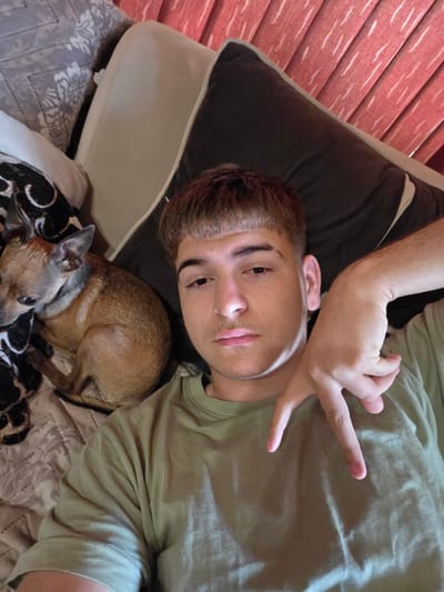 The image shows a close-up, overhead view of a young male and a small dog lying on a couch. The male, with short brown hair and prominent eyebrows, is wearing a light green t-shirt and is posing with his hand making a 'V' sign. He is looking directly at the camera. A small, light brown chihuahua is curled up to his left, resting on a patterned blanket. The background of the image consists of pillows and what appears to be blinds or a textured wall. The lighting suggests an indoor environment. There is no discernible information in the image that would place the scene in Aleksandrovo, Bulgaria.