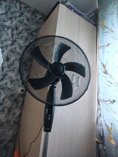 The image shows a black oscillating fan with a metal grate and black blades. The fan is standing on a metal pole and is positioned in front of a light-colored wardrobe. To the left of the fan is a wall with a textured gray pattern, and to the right is a wall with a marble-like pattern featuring gold and blue streaks. A black power cord from the fan runs down the pole and plugs into a white electrical outlet on the gray wall. The fan is the primary object in focus. The scene appears to be indoors, possibly a room in a home or apartment. There are no people or animals visible in the image. The lighting suggests it could be daytime, but without external windows or clocks, it's impossible to determine the time of day or weather. No specific emotions are conveyed, and there is no visible text other than a small logo on the fan's pole which is too small to read. The location context provided is Artemovsky, Russia, but there are no visual cues in the image that specifically indicate this location.
