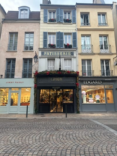 The image displays a row of three multi-story buildings, primarily residential above ground-floor commercial establishments, situated on a cobblestone street in Saint-Germain-en-Laye, France, under an overcast sky.
From left to right:
1. A light beige four-story building with a dormer window on its roof. Its windows have light grey frames and narrow balconies with railings. The ground floor houses "JIMMY FAIRLY," an optical store with a light mint-green facade, brightly illuminated interior, and displays of eyeglasses.
2. A central, cream-colored four-story building. Its upper-floor windows are fitted with light blue louvered shutters and decorated with small red bows and greenery. A gold-lettered sign, "PATISSERIE," is affixed above the ground floor. The ground-floor shop is "GRANDIN," identified by a dark grey facade, a black awning, and extensive festive green garlands with red bows and integrated lights. The brightly lit interior reveals display cases with baked goods. One individual is visible inside, facing away from the camera, wearing light-colored clothing and a yellow-green backpack.
3. A light yellow four-story building. Its windows feature black ornate wrought-iron balconies; one window on the second floor is partially open. The ground floor contains "EDGARD OPTICIENS," an optical store with a grey facade and a window display featuring several large, pastel-colored, abstract sculptural objects.
The street surface in the foreground is paved with light brown and grey cobblestones arranged in a radiating, fan-like pattern. Three dark grey cylindrical bollards are positioned along the curb in front of the "JIMMY FAIRLY" and "GRANDIN" shops. A black street lamp, featuring a spherical lantern, is mounted between the left and central buildings.