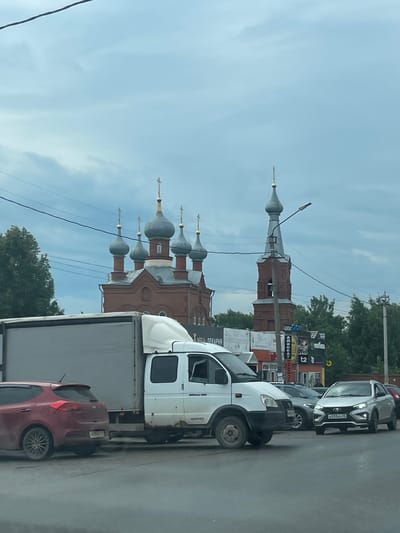 The image depicts a street scene in Malaya Purga, Russia, characterized by a line of parked vehicles. In the foreground, a white truck with a grey trailer is prominent, followed by a red car. Behind them, other vehicles, including a dark grey SUV and a lighter grey car, are also visible.

The backdrop features a notable architectural element: a brick church with multiple domed towers, suggesting a historical or religious site. The sky is overcast with clouds, indicating a possibly humid or temperate weather condition. Trees line the background, interspersed with utility poles and wires, common in urban or suburban environments. Several signs with Cyrillic text are present, though their content is not entirely legible. One sign appears to say "АВТОКАТ". Another sign, partially obscured, seems to indicate a business name ending in "ДЕКАРНА".

The overall impression is that of a typical daytime street scene, possibly during rush hour or a period of moderate traffic, given the parked vehicles. The presence of the church adds a cultural and historical layer to the otherwise ordinary urban landscape. No people are directly visible in the image.