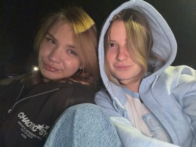 Two young women are shown in a close-up, selfie-style photograph taken at night. The woman on the left has light brown hair and is wearing a dark-colored zip-up hoodie with white text and graphics, and light blue jeans. The woman on the right has blonde hair and is wearing a light blue hooded sweatshirt over a light-colored sweater with a patterned design. They are seated, likely in a vehicle, as evidenced by the background which appears to be dark and the suggestion of a car interior. The lighting suggests artificial light sources. There is no indication of the city of Siedlce, Poland, or any other specific location within the image.