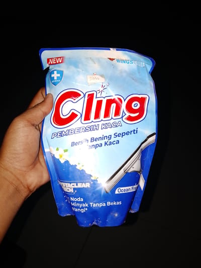 A hand is holding a blue and white pouch of Cling glass cleaner. The pouch has red lettering that says "CLING" and smaller text that says "PEMBERSIH KACA" and "Bersih Bening Seperti Tanpa Kaca". The pouch also features an image of a squeegee and some white flowers. In the top left corner, there is a red and white "NEW" tag and a blue shield with a white plus sign and the word "HIGIENIS". In the top right corner, there is the "WINGS CARE" logo. On the bottom left of the pouch, there is text that says "CRYSTACLEARTTECH" and "Noda Minyak Tanpa Bekas Wangi*". On the bottom right, the pouch indicates "Ocean Fres". The product is being held against a black background, suggesting it is indoors or at night. The image is taken in Sendangrejo, Indonesia.