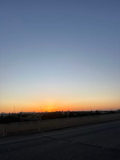 An outdoor scene captured during sunset or sunrise displays an urban horizon from the perspective of a highway. The sky transitions from a deep blue at the zenith to vibrant orange and yellow hues at the horizon, where the sun is partially visible as a bright orange disc. Numerous small, dark specks, consistent with birds, are scattered across the upper and middle portions of the sky.

Along the horizon, a prominent silhouette of a city skyline is visible, characterized by multiple buildings of varying heights. Below the skyline and across the mid-ground, a series of streetlights with curved poles extend, some illuminated or reflecting the intense light. Dark vegetation, likely trees and bushes, forms a band beneath the urban structures. Further to the right in the mid-ground, an indistinct area possibly indicates a parking lot or industrial zone.

In the foreground, a dark asphalt road occupies the bottom of the frame, angled slightly from left to right. It exhibits subtle cracking and faint linear markings. A light-colored concrete barrier or retaining wall separates the road from the distant urban landscape, running horizontally across the image. No individuals, specific actions, or interactions are depicted. The scene is situated within the geographical area of Sdot Dan Regional Council, Israel, with the view extending towards an urban center.