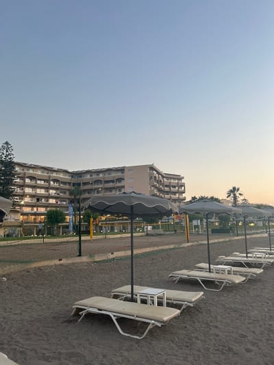 The image depicts a beach scene on the island of Rhodes, Greece, likely in the late afternoon or early evening as indicated by the soft lighting. In the background, a large, multi-story hotel with numerous balconies and windows is visible. The hotel is set back from the beach, with greenery and palm trees in front of it.
The foreground of the image is dominated by a sandy beach. Arranged in rows are several empty beach loungers, most of which are a light beige color with white frames. Small, white side tables are placed between some of the loungers. Overlapping these loungers are several large, gray beach umbrellas, providing shade.
Further back on the beach, there are two volleyball courts delineated by nets and poles. The sand in the court areas appears to be lighter in color than the sand in the foreground. Some people are visible in the distance, near the hotel and the volleyball courts, though they are too small to discern specific details. A volleyball net is strung across one of the courts, with yellow poles supporting it. The overall atmosphere is one of a resort or leisure area, prepared for visitors but currently not crowded. The sky above is a clear, soft gradient of blue, transitioning to a paler hue near the horizon.
