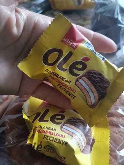 In Altinkul, Uzbekistan, a close-up shot focuses on a person's hand holding two yellow wrappers of "Olé" brand chocolates. The wrappers display the brand name in large, brown lettering and an illustration of a chocolate-covered biscuit with a white marshmallow filling. Text on the wrappers includes "KREMBER," "GLAZURLANGAN," "MARSHMEOULLI," and "PECHENYE." The background is blurred, showing hints of other food items and packaging. The lighting is natural, suggesting an indoor setting.