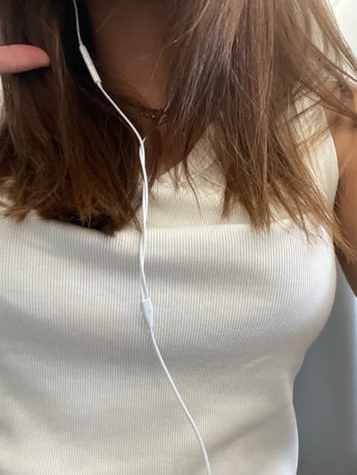 This close-up image, attributed to Villeneuve-le-Roi, France, depicts the upper torso and head of an individual, presumed female, from an eye-level perspective. The subject has medium-brown hair, styled to fall around the shoulders and neck. She wears a white, vertically ribbed tank top. A thin silver chain necklace is visible around her neck, partially obscured by hair. White wired earphones are in use; the cord descends vertically over the sternum, featuring a small white splitter. The tip of a finger is briefly visible near the left temple, partially obscured by hair. The immediate background on the right consists of a plain, light-colored surface. No discernible actions beyond wearing the depicted items are observed.