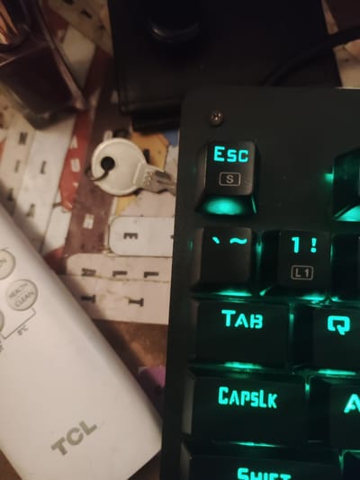 The image shows a close-up view of a mechanical keyboard with illuminated green keys. The keys visible include "ESC," "S," "~", "1!", "TAB," "CAPSLK," and parts of other keys. In the background, a silver key with the number "886" on it rests on what appears to be a printed surface. To the left of the keyboard, a white TCL remote control is partially visible, with buttons labeled "EN," "HEALTH," and "CLEAN." The overall scene is an indoor setting, likely a desk or table, with the background out of focus. There is no indication of the location being Roranwala, Pakistan, or any other specific city. The image focuses on the keyboard and the surrounding objects.