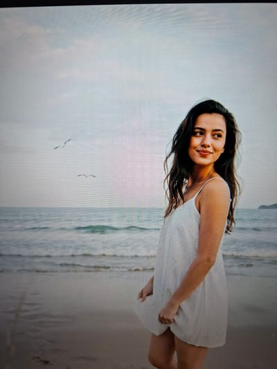 A young woman with long, dark hair is captured in a medium shot, seemingly in motion on a beach. She is wearing a white, sleeveless dress. Her head is tilted back slightly, and she is looking towards the viewer with a soft smile. The background is dominated by a hazy, overcast sky and the ocean with gentle waves. The wet sand of the beach is visible in the foreground. Two small birds are flying in the distance over the water. The lighting suggests it could be overcast or late in the day. There is no visible text in the image.