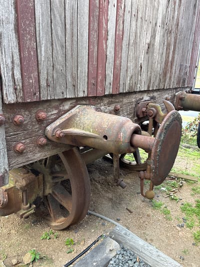 The image shows a section of what appears to be an antique or abandoned piece of machinery resting on a weathered, wooden platform. The machinery includes a rusty seat attached to a vertical pipe, supported by a base fixed to the wooden platform with several large bolts. The platform has peeling paint, revealing layers of white and red hues on the wood. A large rusty wheel is visible next to the base of the machinery. The ground adjacent to the platform is a mixture of dirt and small rocks with some sparse vegetation. The location is Marindia, Uruguay, but there are no people, actions, or interactions present in the frame.